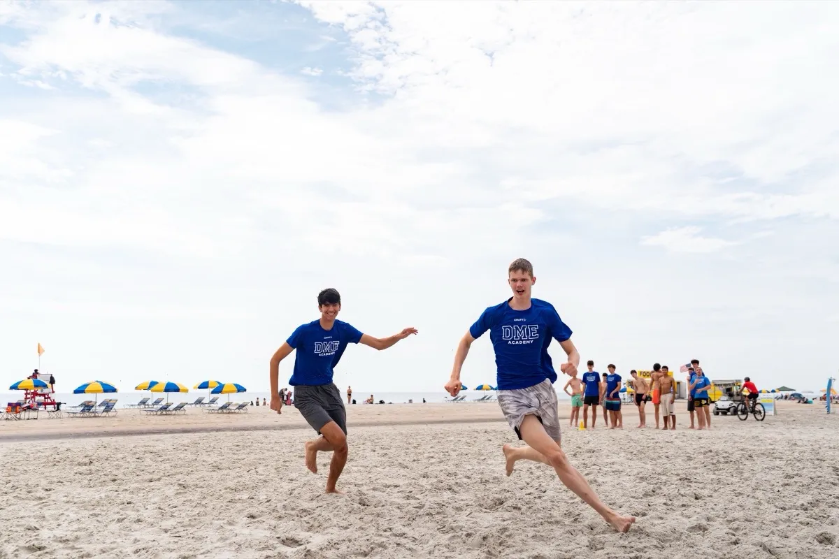 Two athletes running barefoot on beach in DME Academy shirts
